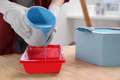Woman pouring light blue paint from bucket into tray at wooden table indoors, closeup