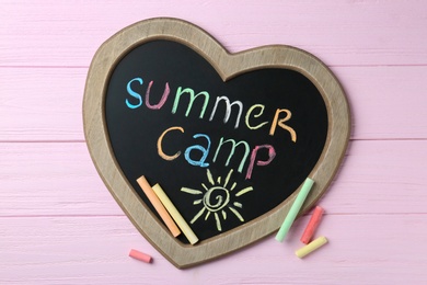 Photo of Heart shaped blackboard with text SUMMER CAMP, drawing and chalk sticks on wooden background, flat lay