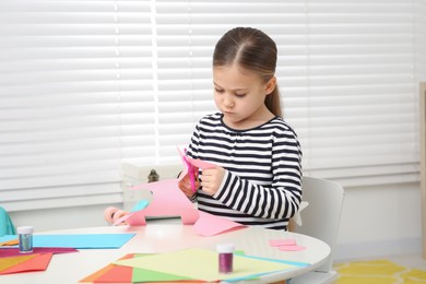 Photo of Cute little girl cutting pink paper at desk in room. Home workplace