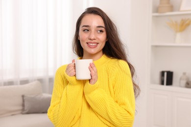 Photo of Beautiful young woman in stylish warm sweater holding cup of drink at home