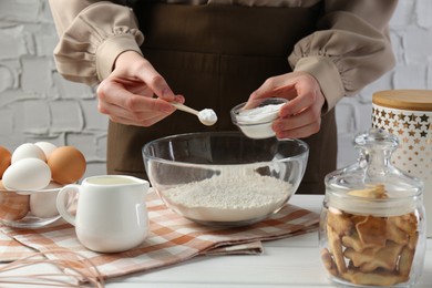 Photo of Woman with spoon and bowl of baking powder at white wooden table, closeup