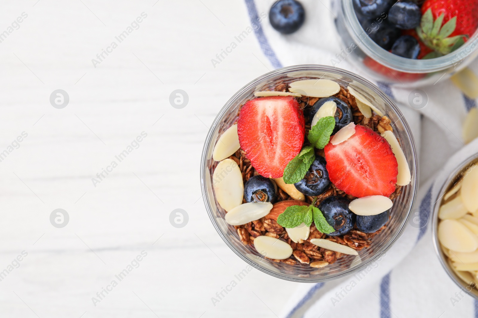 Photo of Tasty granola with berries, almond flakes and mint in glass on white table, flat lay. Space for text