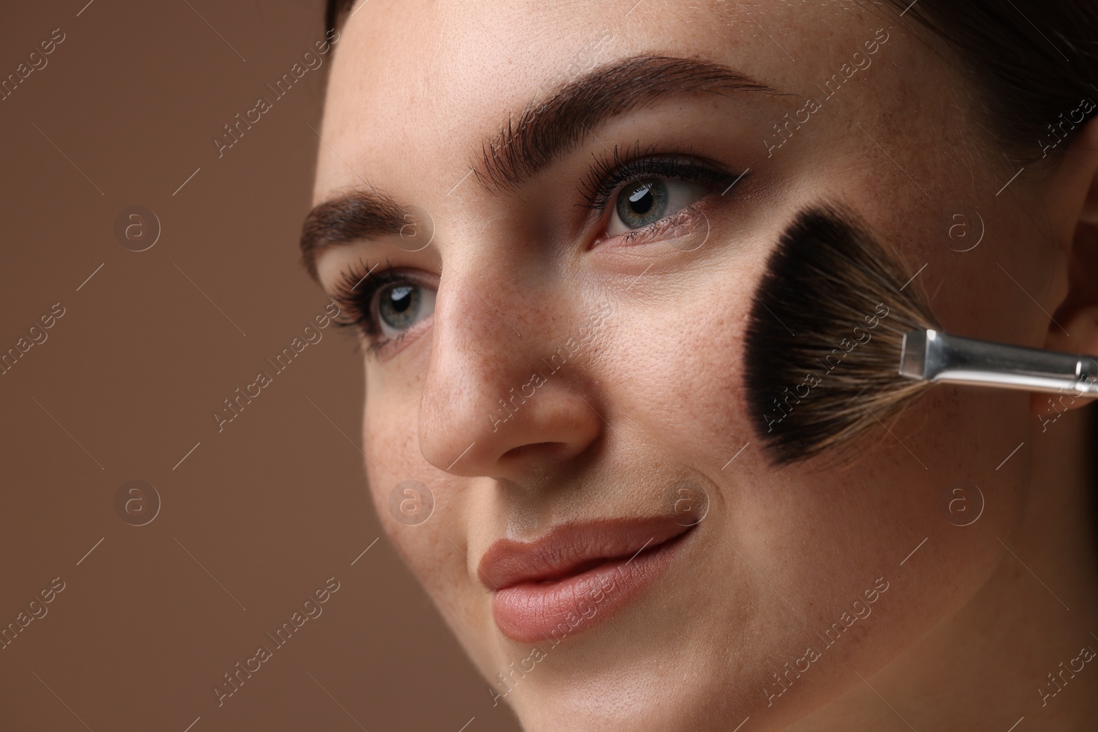 Photo of Beautiful woman with freckles applying makeup with brush on brown background, closeup