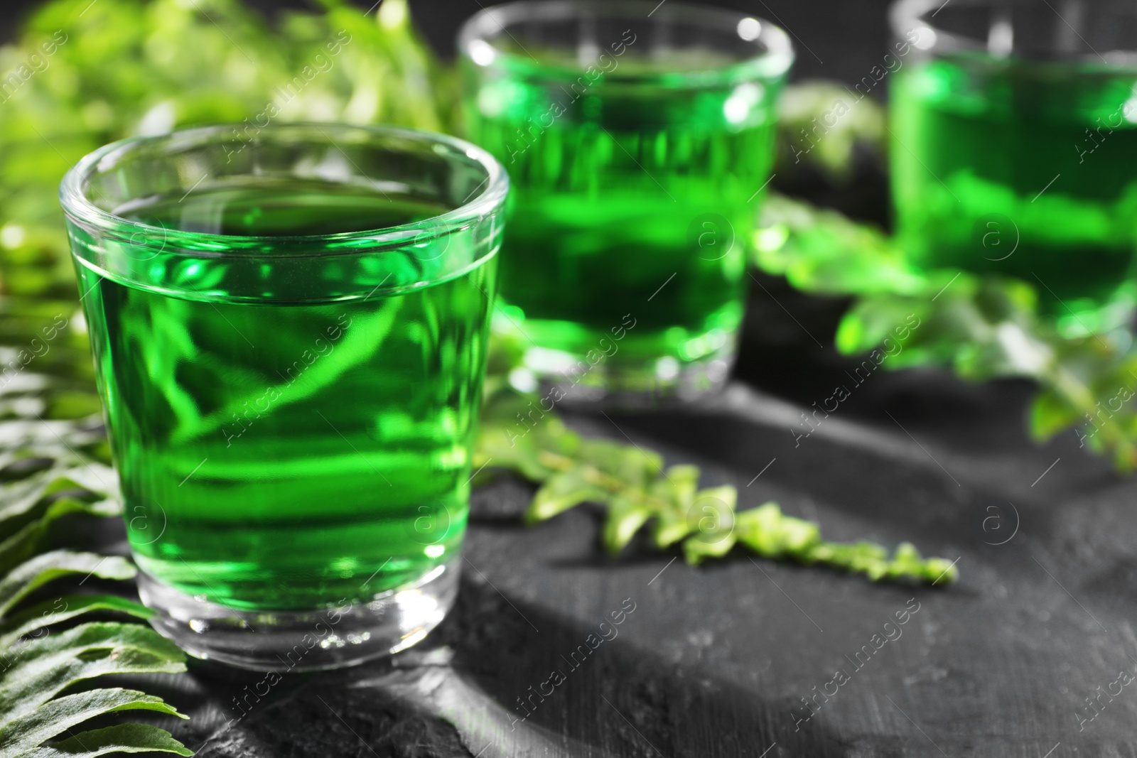 Photo of Absinthe in shot glasses and green leaves on black table, space for text. Alcoholic drink