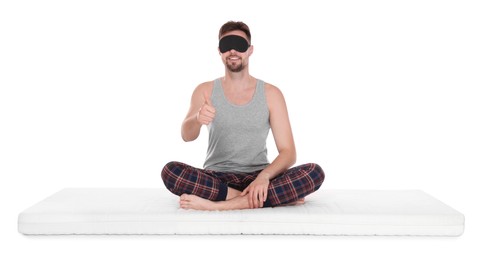 Photo of Smiling man in sleeping mask sitting on soft mattress and showing thumb up against white background