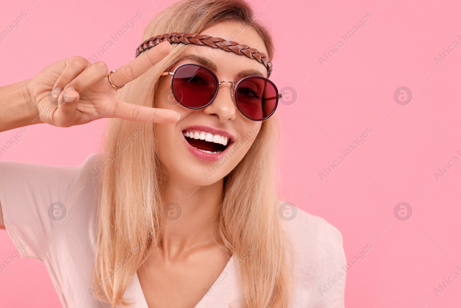 Photo of Portrait of smiling hippie woman showing peace sign on pink background. Space for text