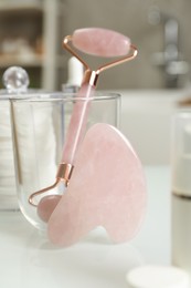 Rose quartz gua sha tool, natural face roller and toiletries on white table indoors