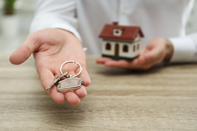 Photo of Real estate agent holding house key with trinket at wooden table, closeup