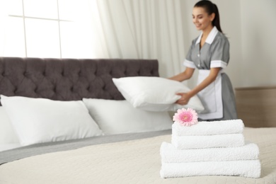 Photo of Stack of clean towels with flower on bed and blurred chambermaid in hotel room, closeup. Space for text