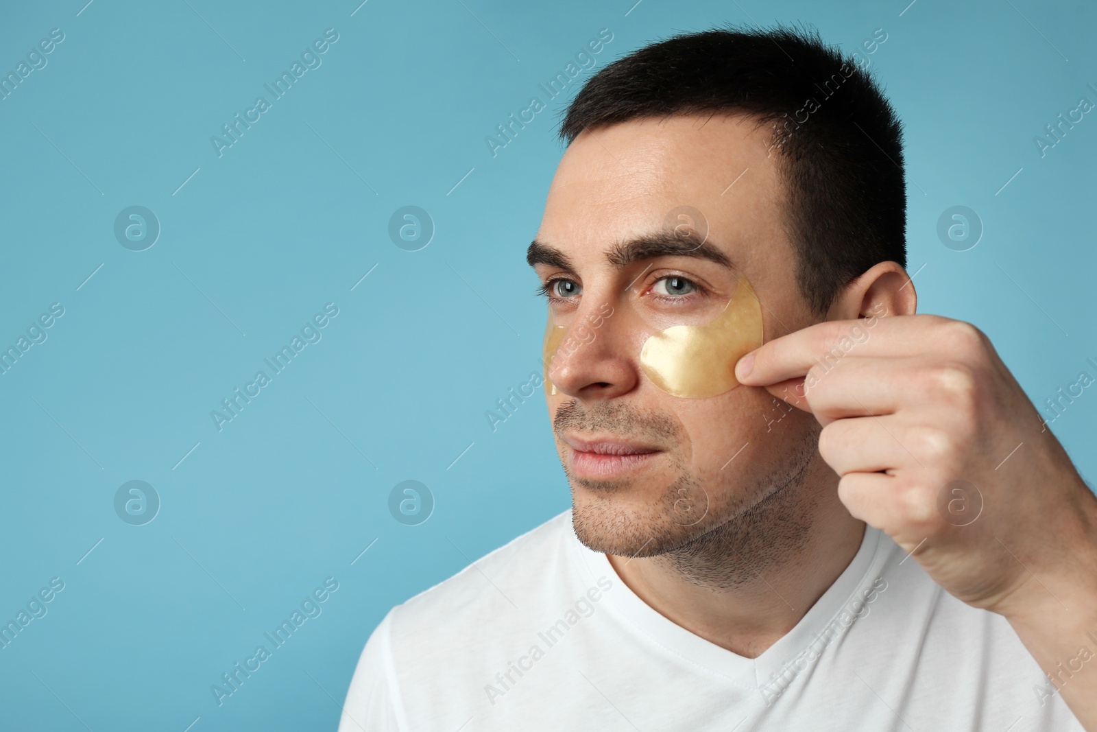 Photo of Young man applying under eye patches on light blue background. Space for text