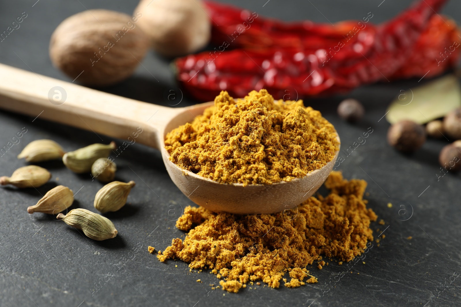 Photo of Spoon with dry curry powder and other spices on dark textured table, closeup