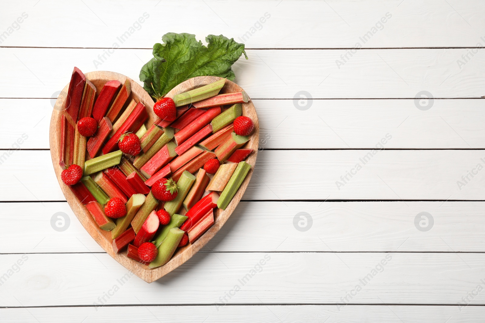 Photo of Cut fresh rhubarb stalks and strawberries on white wooden table, top view. Space for text