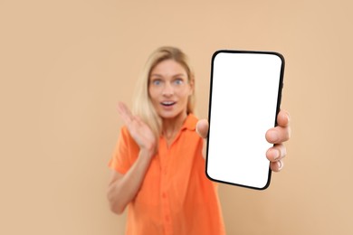 Surprised woman holding smartphone with blank screen on beige background, selective focus