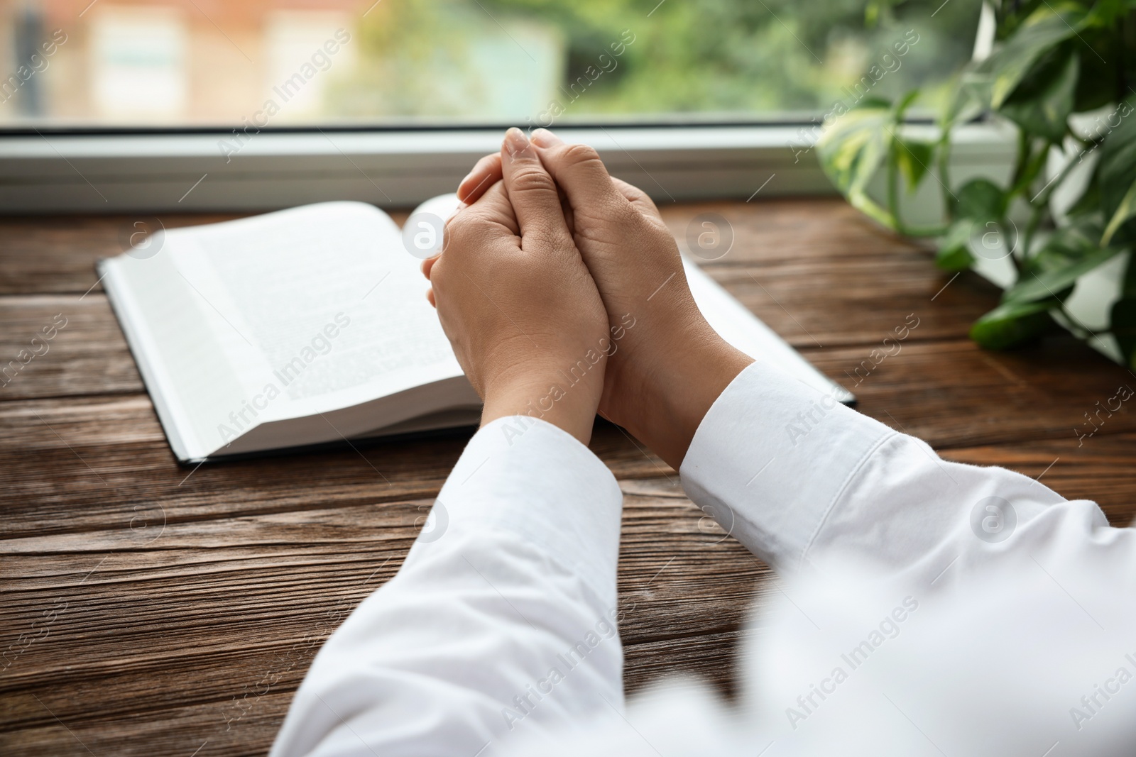 Photo of Woman holding hands clasped while praying at wooden table with Bible, closeup