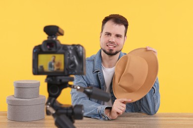 Photo of Smiling fashion blogger showing hat while recording video at table against orange background