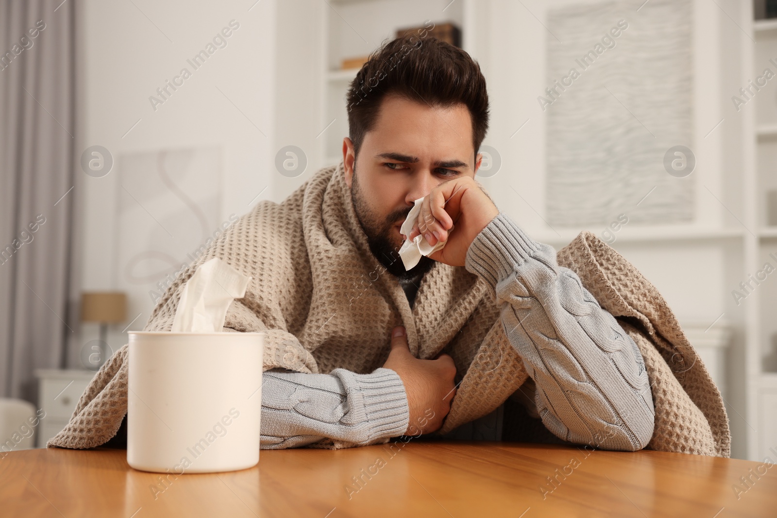 Photo of Sick man wrapped in blanket with tissue blowing nose at wooden table indoors. Cold symptoms