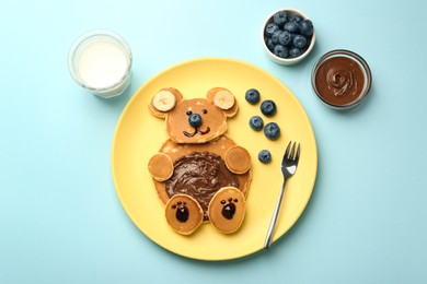 Creative serving for kids. Plate with cute bear made of pancakes, blueberries, bananas and chocolate paste on light blue table, flat lay