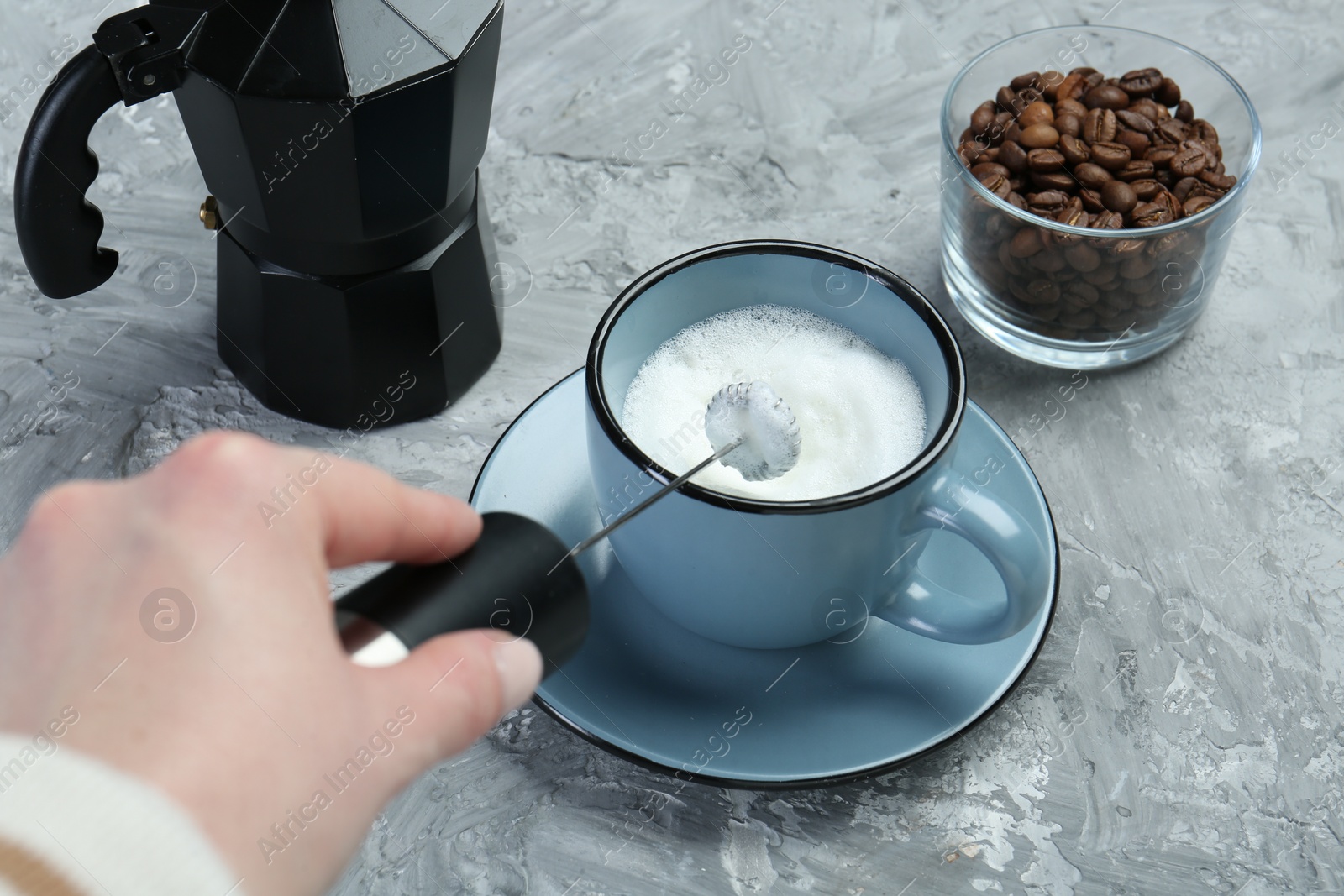 Photo of Woman whisking milk in cup with mini mixer (milk frother) at grey textured table, closeup