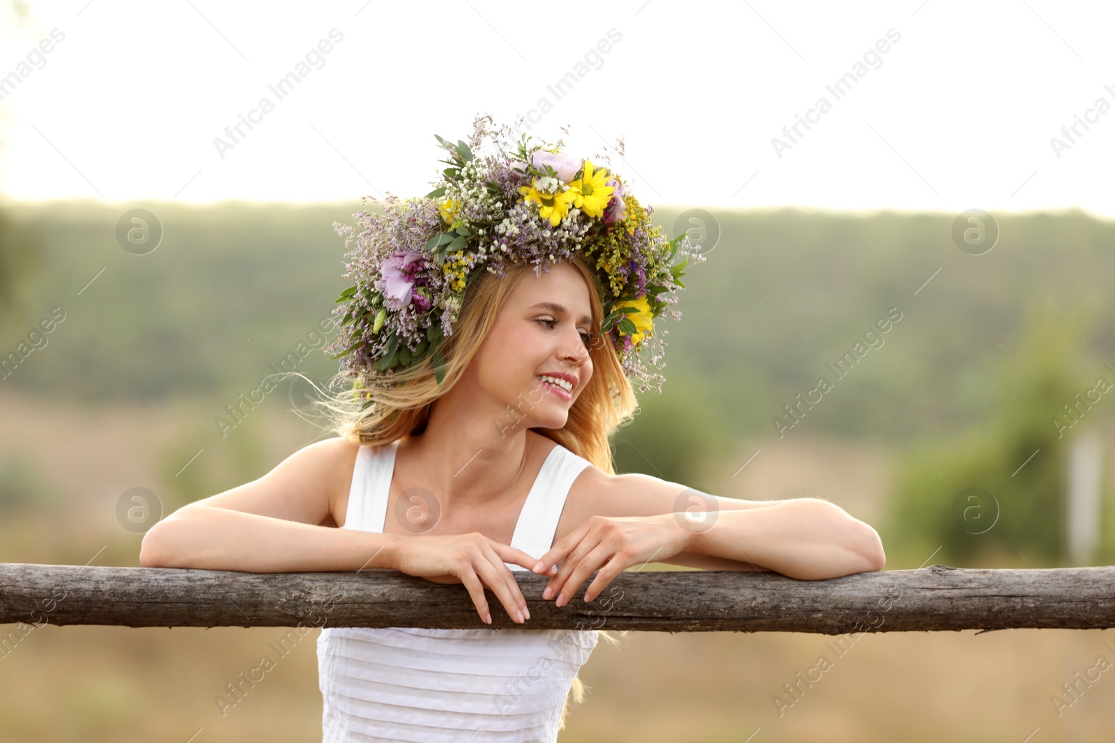 Photo of Young woman wearing wreath made of beautiful flowers near wooden fence