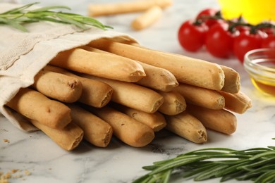 Photo of Delicious grissini sticks, oil, rosemary and tomatoes on white marble table, closeup