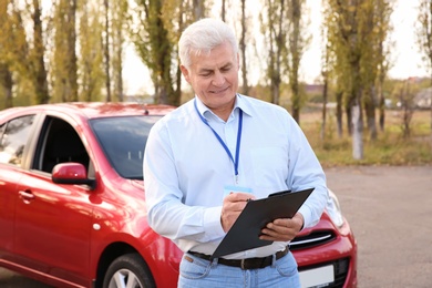 Senior instructor with clipboard near car outdoors. Get driving license