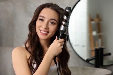 Photo of Smiling woman using curling hair iron in bathroom
