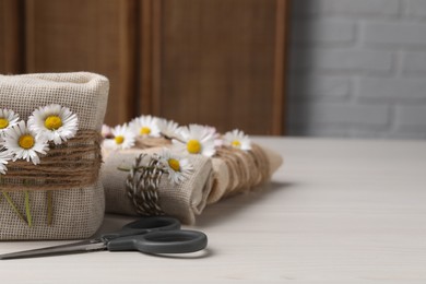 Gifts packed in burlap fabric, beautiful chamomiles, ropes and scissors on white table. Space for text