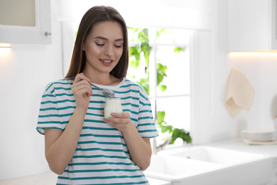 Young attractive woman eating tasty yogurt in kitchen. Space for text
