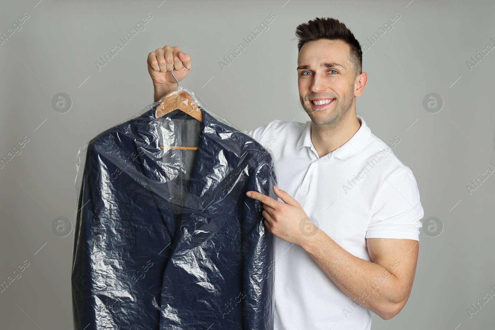 Photo of Man holding hanger with jacket in plastic bag on light grey background. Dry-cleaning service