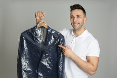 Photo of Man holding hanger with jacket in plastic bag on light grey background. Dry-cleaning service