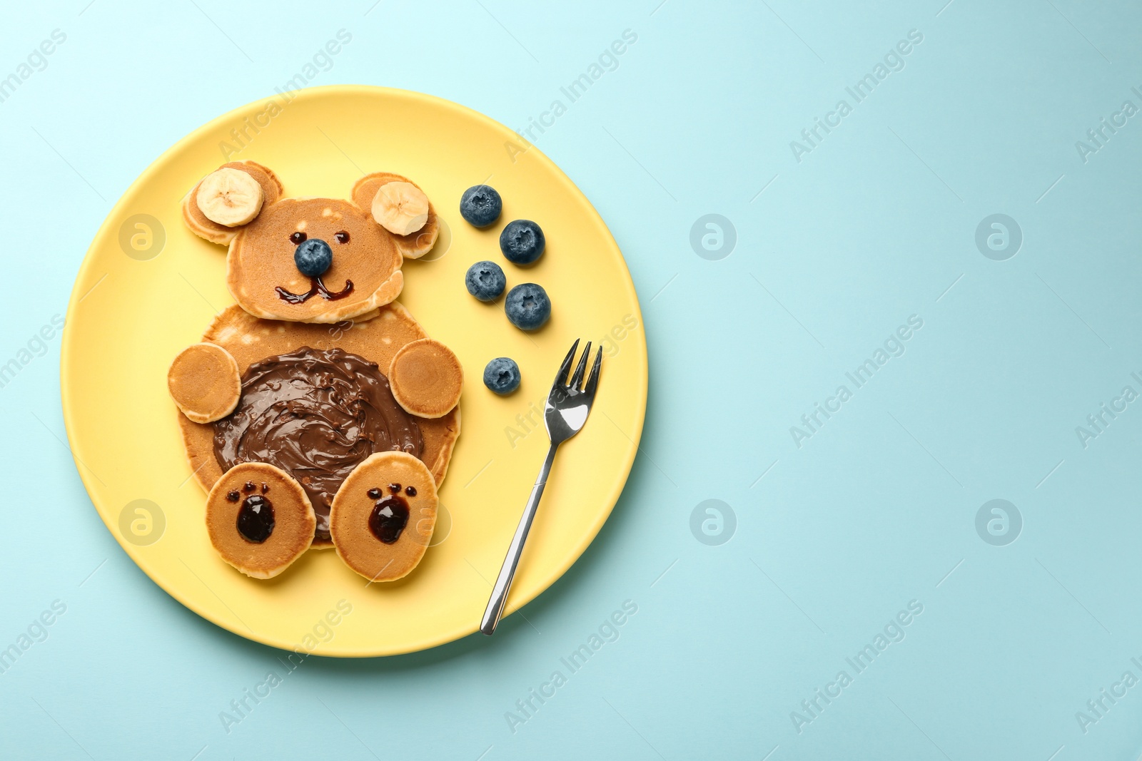 Photo of Creative serving for kids. Plate with cute bear made of pancakes, blueberries, bananas and chocolate paste on light blue table, top view. Space for text