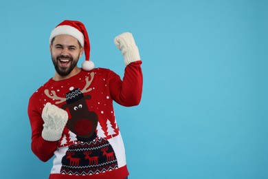 Photo of Happy young man in Christmas sweater, Santa hat on and knitted mittens against light blue background. Space for text