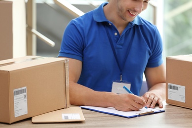 Photo of Young courier working with papers among parcels at table in delivery department