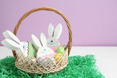 Photo of Wooden bunnies with protective masks, painted eggs in basket and paper grass on white wooden table. Easter holiday during COVID-19 quarantine