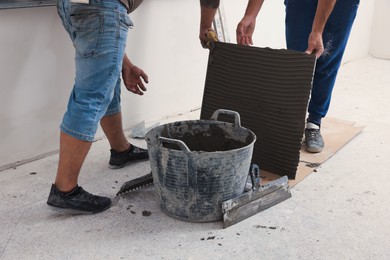 Photo of Workers preparing tile with adhesive mix for installation indoors, closeup