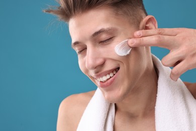 Handsome man applying moisturizing cream onto his face on blue background, closeup