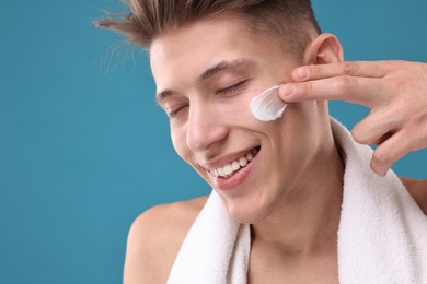 Photo of Handsome man applying moisturizing cream onto his face on blue background, closeup