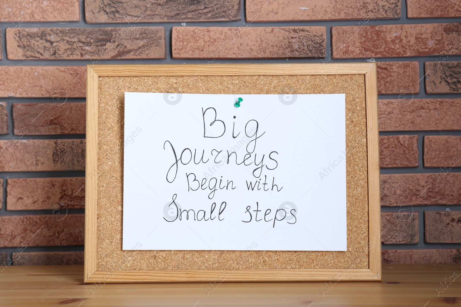 Photo of Corkboard and phrase Big Journeys Begin With Small Steps on table against brick wall. Motivational quote