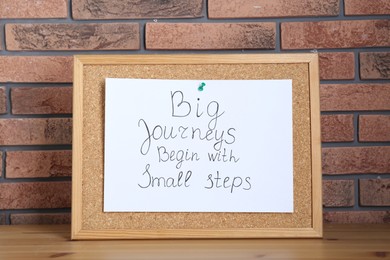 Photo of Corkboard and phrase Big Journeys Begin With Small Steps on table against brick wall. Motivational quote