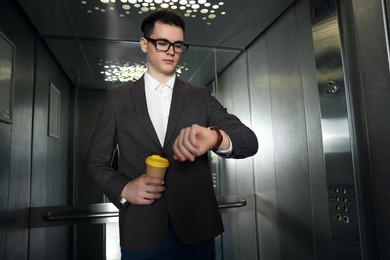 Photo of Young businessman with cup of hot drink checking time in elevator