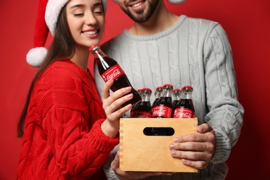 Photo of MYKOLAIV, UKRAINE - JANUARY 27, 2021: Young couple in Christmas hats holding crate with bottles of Coca-Cola on red background, closeup