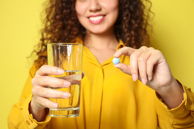 Photo of African-American woman with glass of water and vitamin pill against yellow background, focus on hands