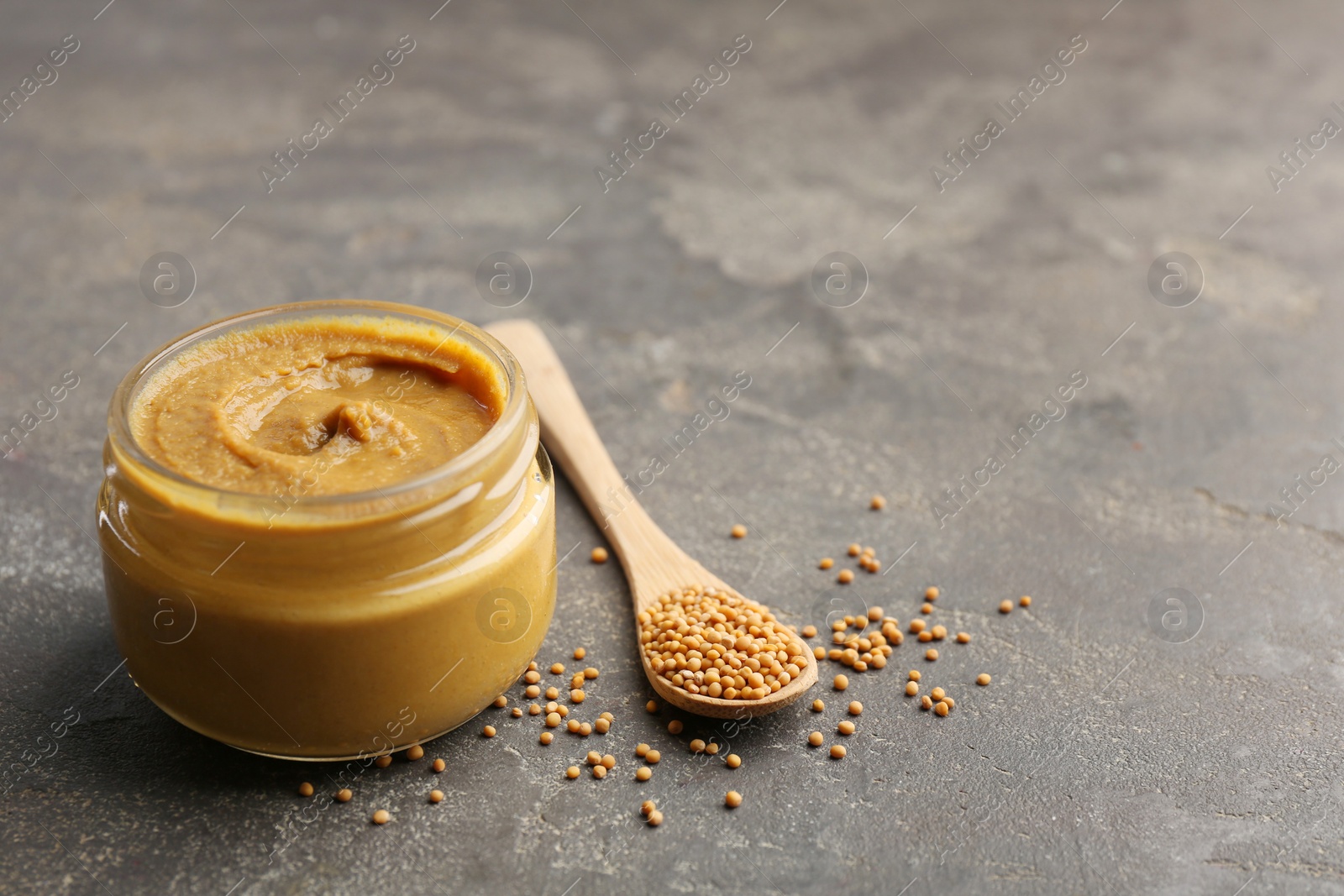 Photo of Tasty mustard sauce in jar and spoon with dry seeds on grey textured table, space for text