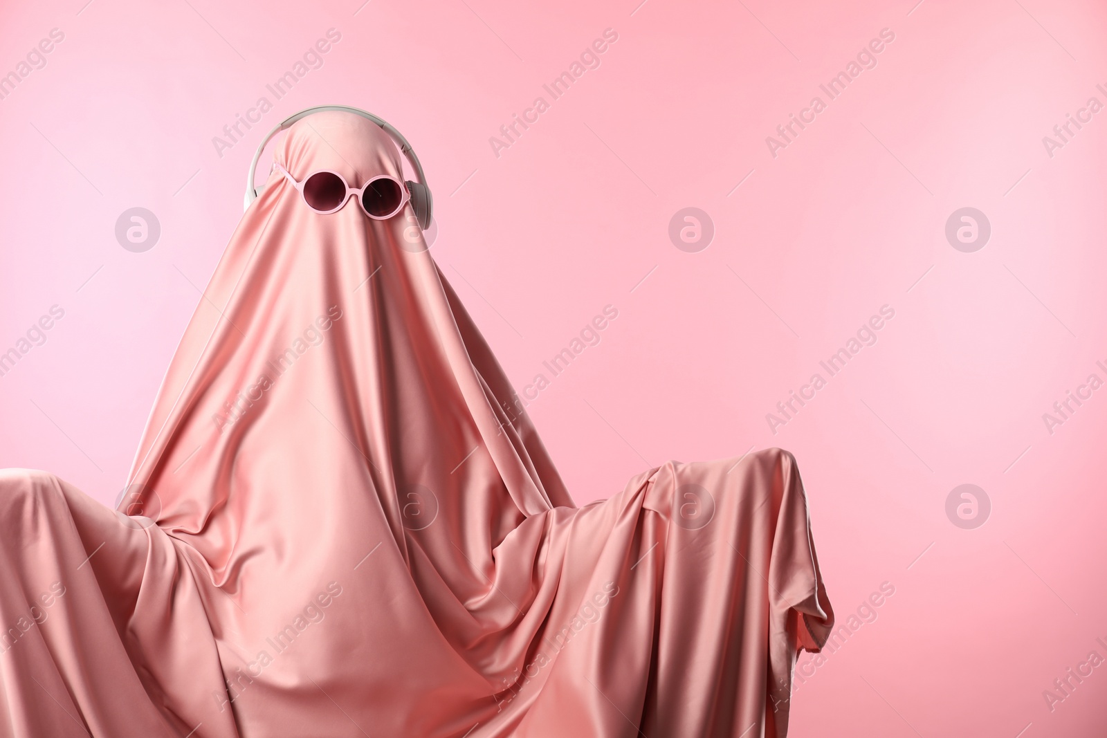 Photo of Glamorous ghost. Woman in sheet with sunglasses and headphones on pink background, space for text