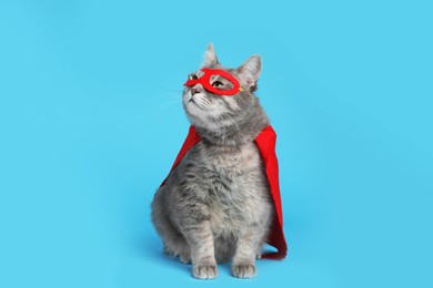 Photo of Adorable cat in red superhero cape and mask on light blue background