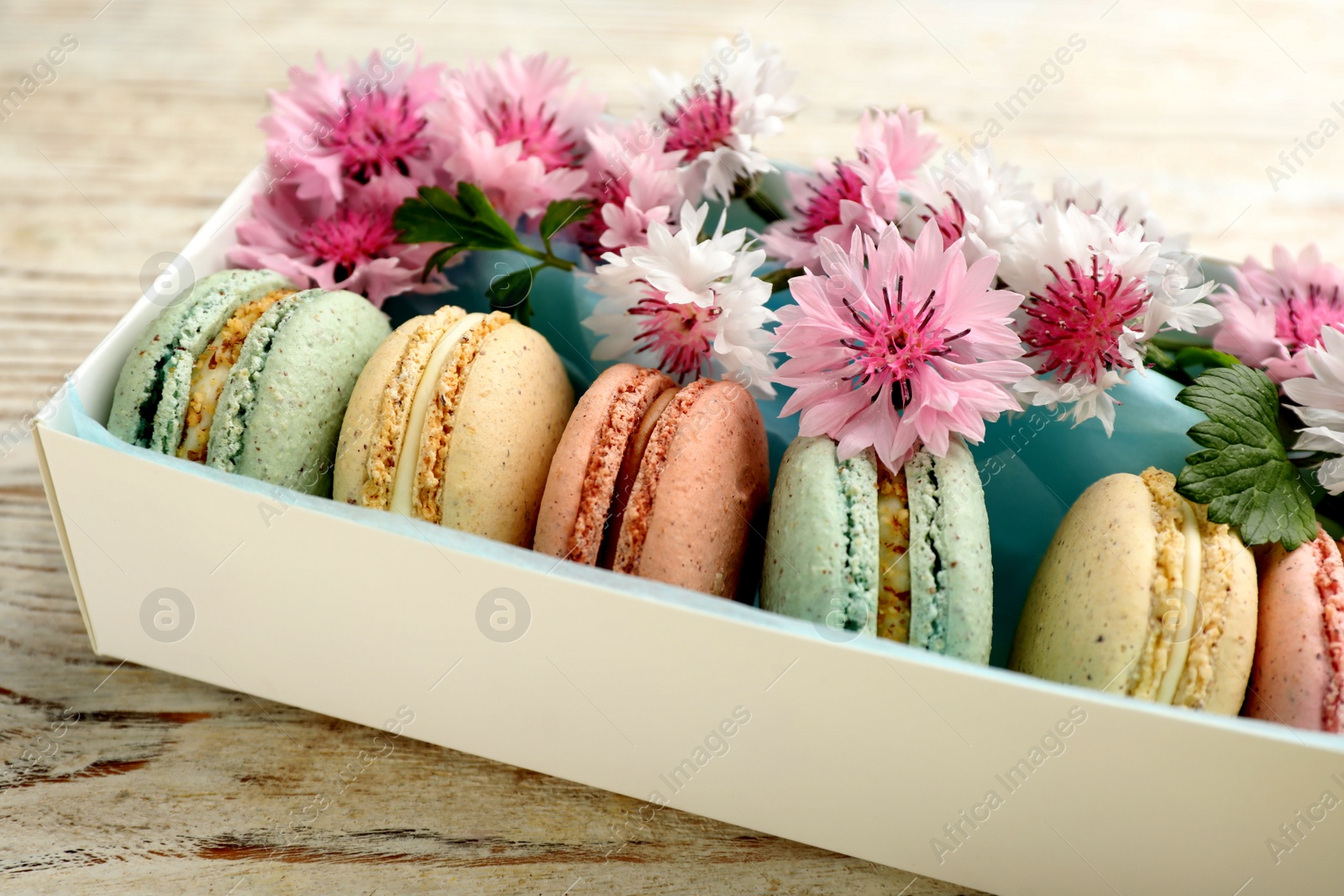 Photo of Delicious macarons and flowers in box on white wooden table