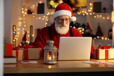 Photo of Santa Claus using laptop at his workplace in room decorated for Christmas