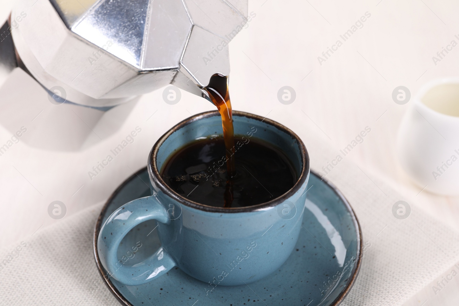 Photo of Pouring aromatic coffee from moka pot into cup at white table, closeup