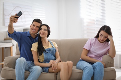 Unhappy woman feeling jealous while couple taking selfie together on sofa at home