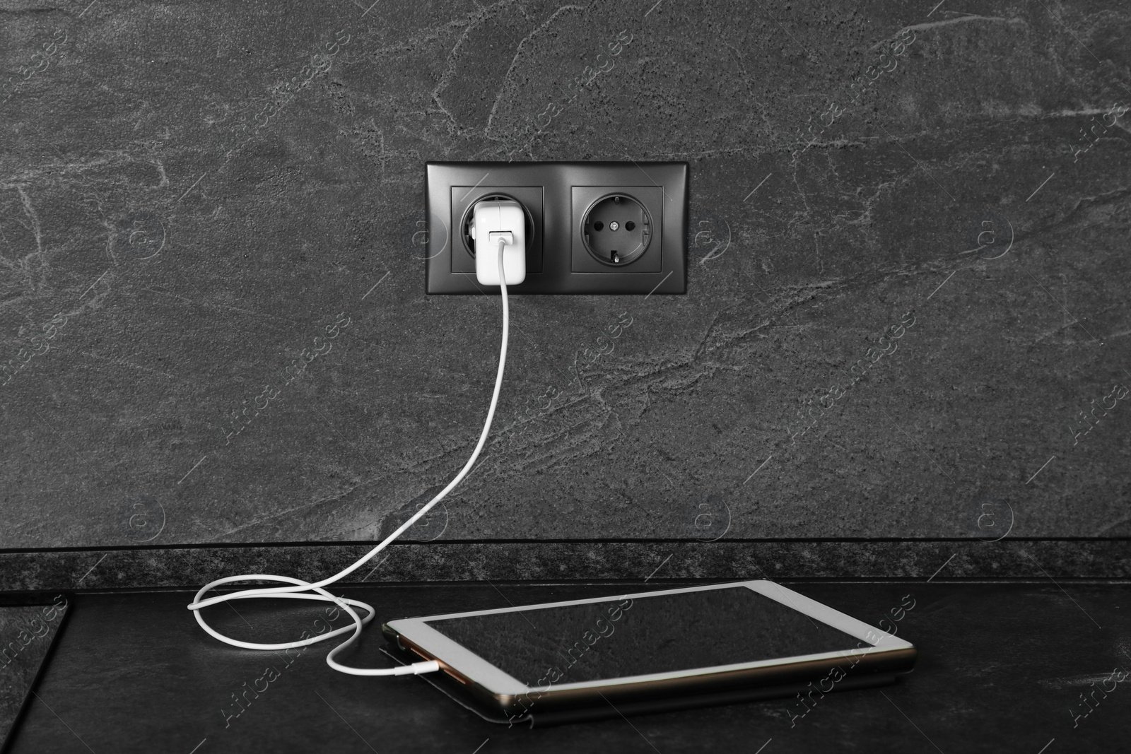 Photo of Tablet charging from electric socket on dark grey table indoors